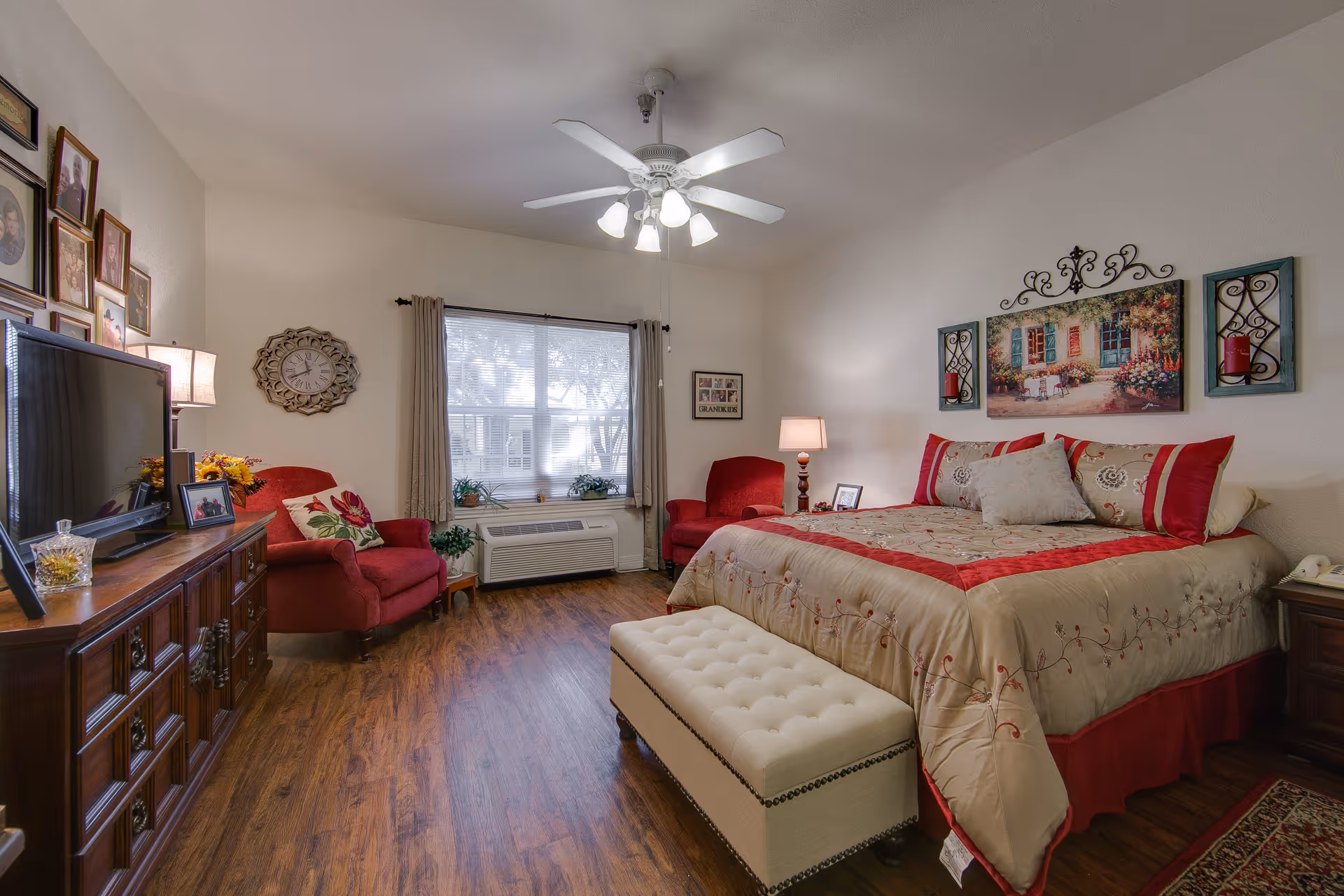 A cozy bedroom with a large bed adorned with beige and red bedding. There is a cushioned bench at the foot of the bed, two red armchairs near a window with beige curtains, a wooden dresser with a TV on top, and framed pictures on the walls. The room has wooden flooring and a ceiling fan with lights.