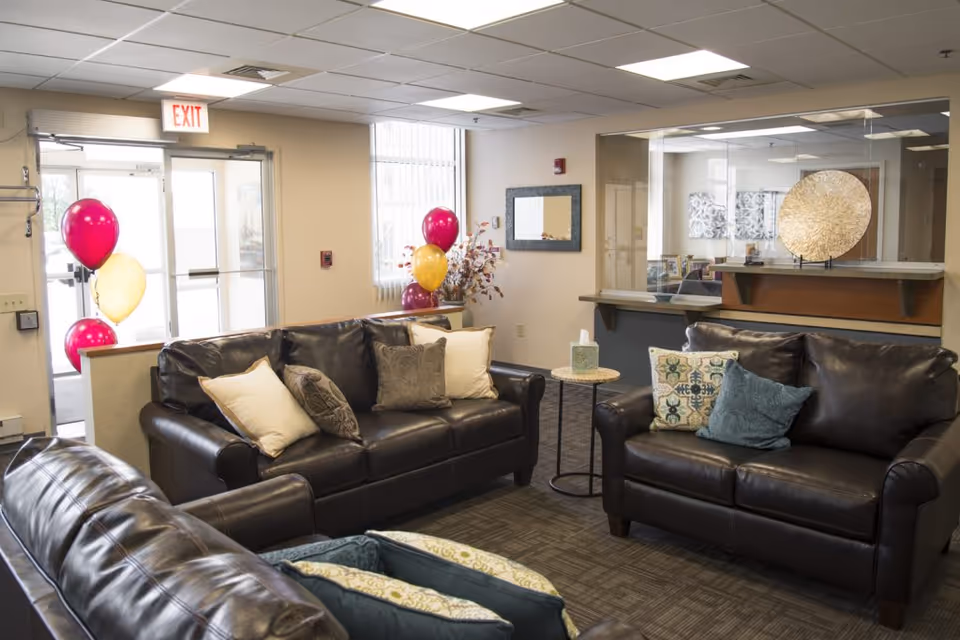 A cozy living room area with three dark leather sofas arranged around a small round side table with a tissue box. The room has a window with vertical blinds, a mirror on the wall, and a decorative golden circular piece on a shelf behind a glass partition. There are colorful balloons near the exit door, and the ceiling has recessed lighting.