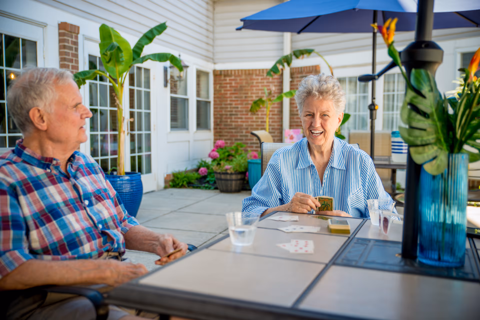 Two elderly people sitting at an outdoor table playing cards. The woman is smiling and holding cards, while the man looks on. The setting is a patio area with potted plants and a building with large windows in the background.