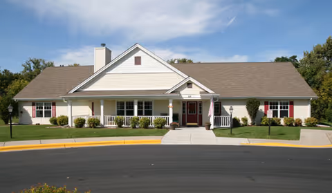 Front exterior view of a single-story building with a gable roof, white siding, and red window shutters. There is a covered entrance with white columns and a walkway leading to the door. The building is surrounded by green grass, bushes, and trees under a partly cloudy sky.