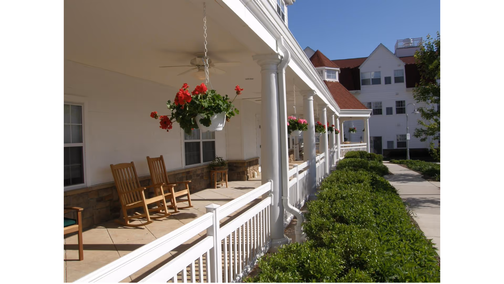 A covered porch area with white columns and hanging flower pots with red and pink flowers. There are wooden rocking chairs on the porch and a walkway lined with green bushes leading to a white building with red roofs in the background.