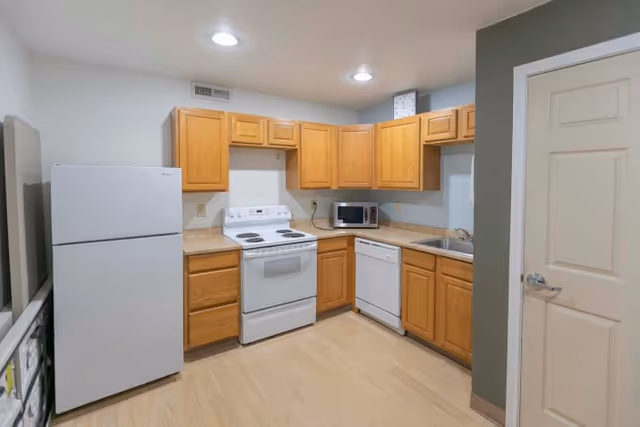 A clean kitchen with light wood cabinets, a white refrigerator, white electric stove, white dishwasher, microwave, and a stainless steel sink. The floor is light wood, and the walls are painted in neutral colors with recessed ceiling lights.