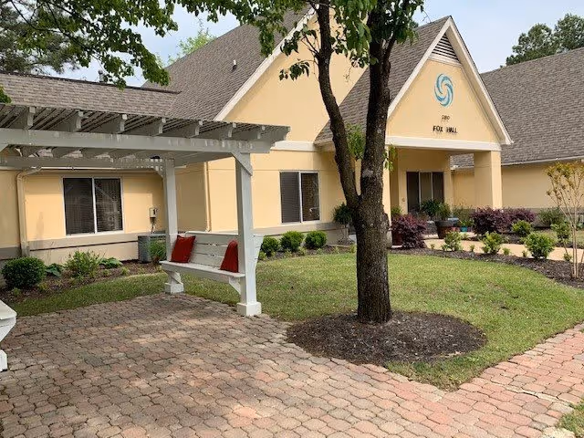 Outdoor courtyard area of a senior living facility named Fox Hall, featuring a paved walkway, a white pergola with a bench that has red cushions, a tree in the center, and a beige building with windows and landscaping in the background.