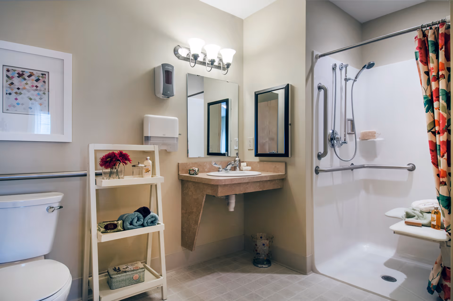 A clean and accessible bathroom with a toilet, a small white shelving unit holding towels and toiletries, a wall-mounted sink with a mirror above it, and a walk-in shower with grab bars and a floral shower curtain.