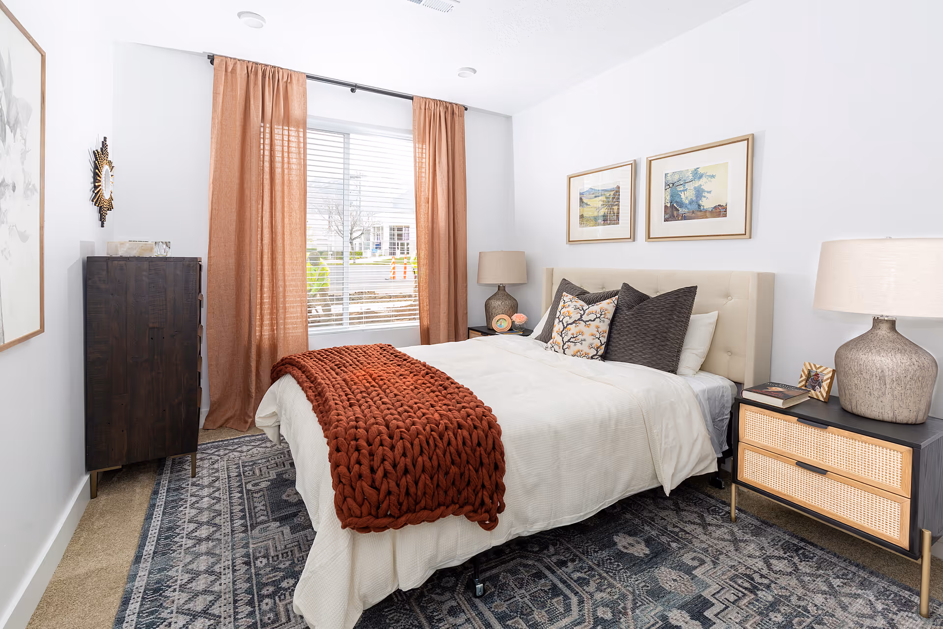 Well-lit bedroom with a bed topped by pillows and a chunky red throw, two bedside tables with lamps, framed art, and a window with rust-colored curtains.