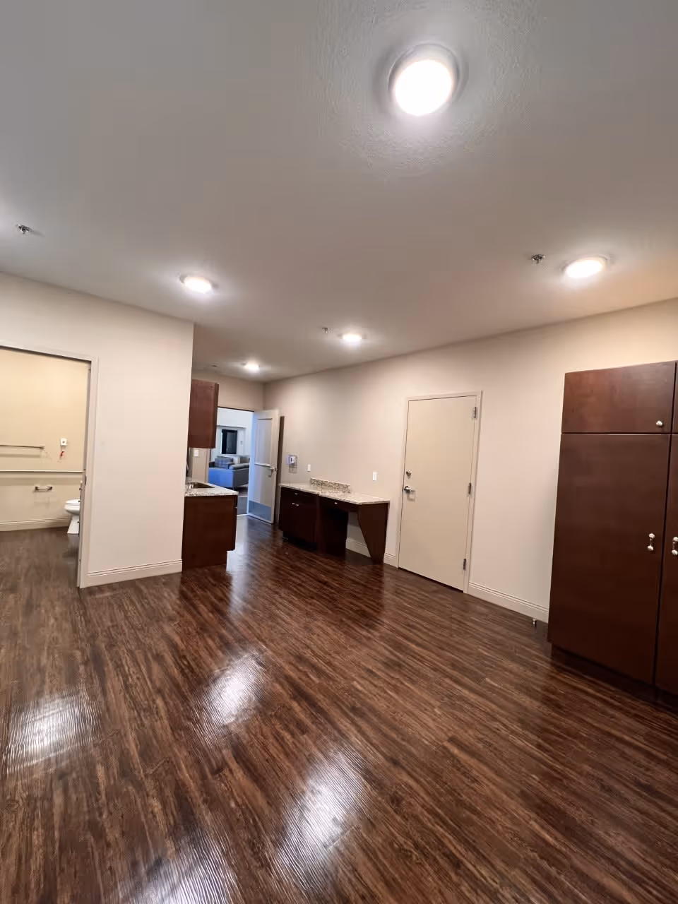 Interior view of a senior living facility room with wood flooring, beige walls, recessed ceiling lights, dark wood cabinets, a small countertop area, and an open door leading to a bathroom with a visible toilet and grab bars.