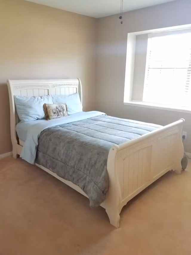 A white ornate bed with blue bedding sits in a beige bedroom with a window.