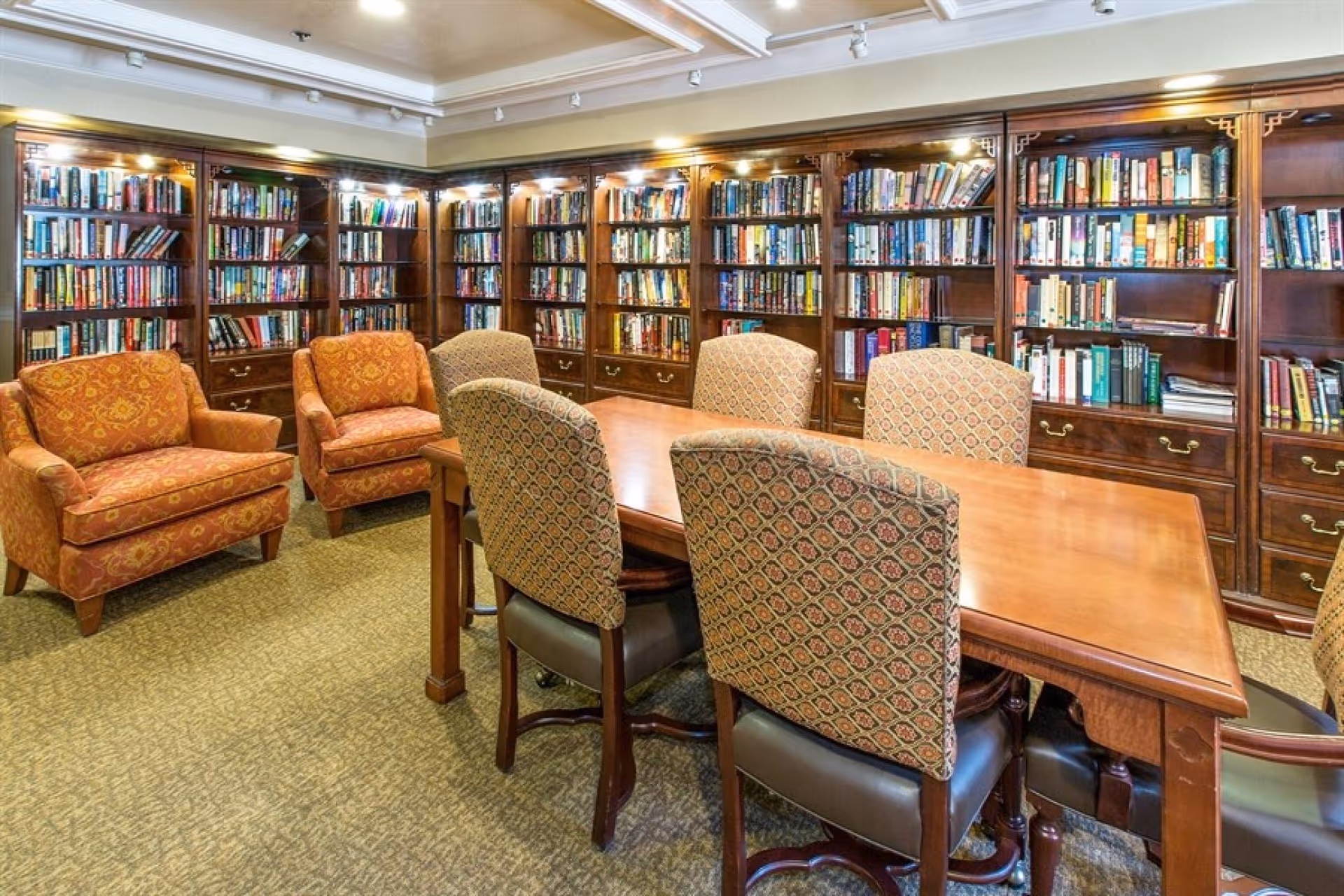 A cozy library/reading room with wooden bookshelves lining the walls, a large wooden table surrounded by upholstered chairs, and patterned armchairs.