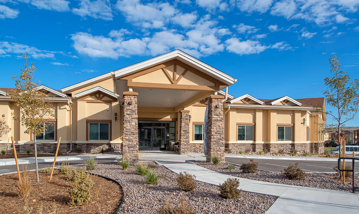 Exterior view of a single-story building with beige walls and stone pillars at the entrance, surrounded by a landscaped area with small trees and shrubs under a blue sky with scattered clouds.
