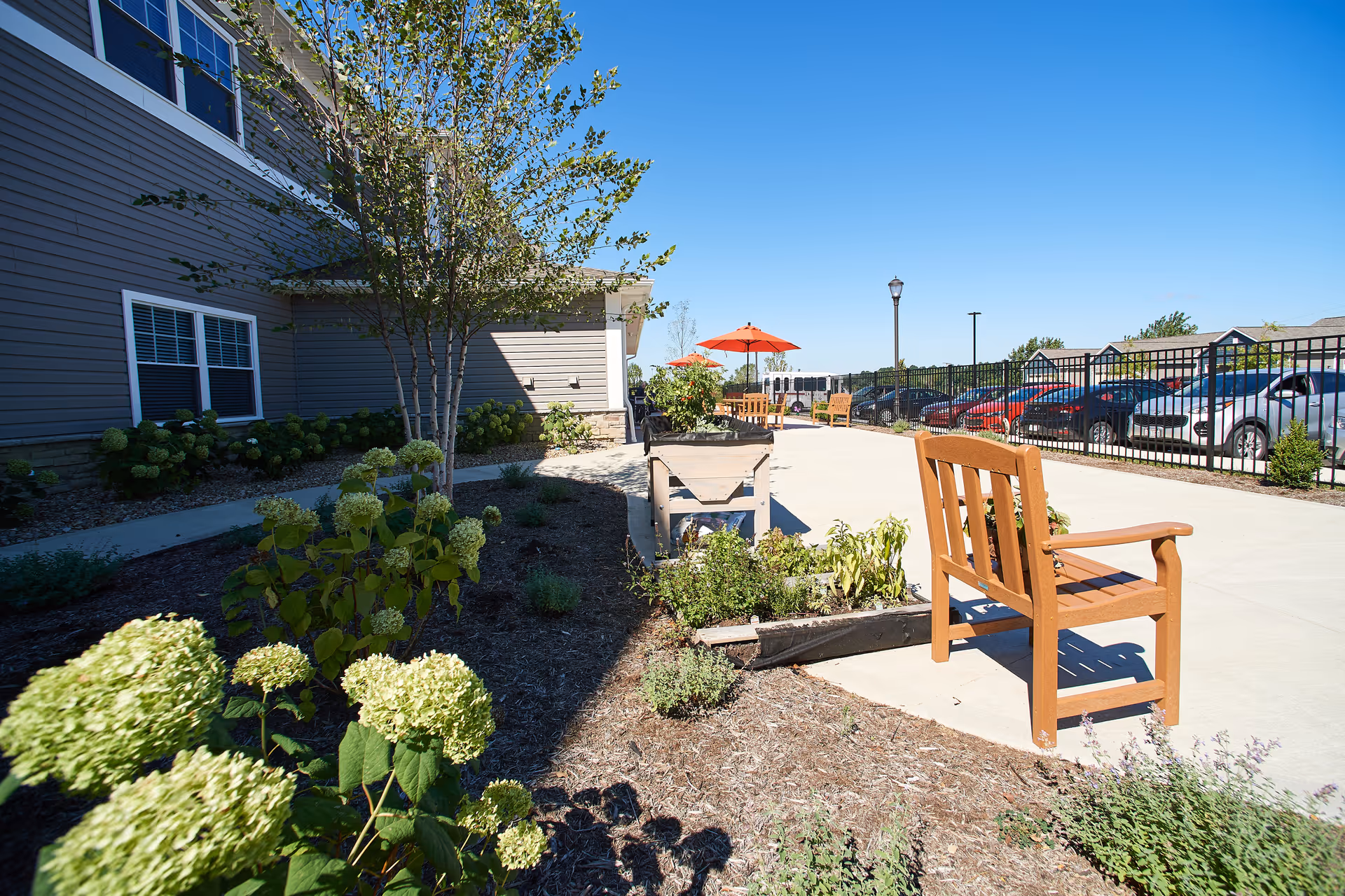 Outdoor courtyard with wooden benches, raised planters, umbrellas and a parking lot beside a building.