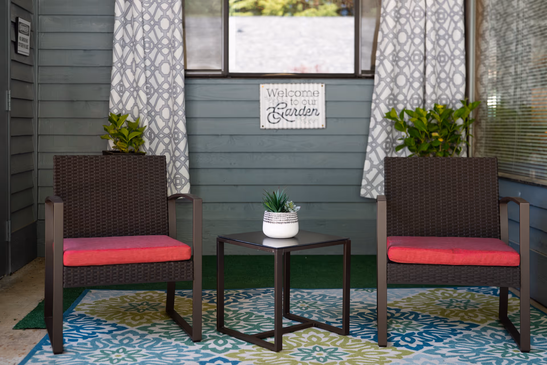 A cozy outdoor seating area with two dark wicker chairs with red cushions, a small black square table with a white potted plant, patterned curtains, green plants on either side, and a sign on the wall that reads 'Welcome to our Garden'.