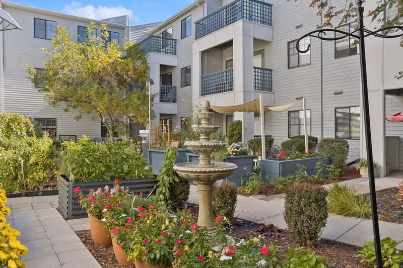 Outdoor garden area at Five Star Premier Residences of Reno featuring a multi-tiered stone fountain surrounded by colorful flowers and plants in pots and raised garden beds, with a three-story residential building in the background under a partly cloudy sky.