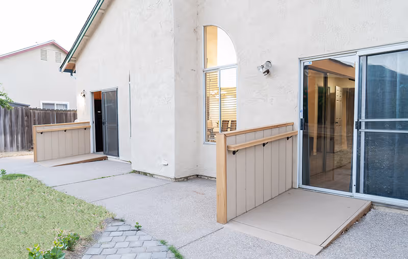 Exterior patio area of a care facility with sliding glass doors, accessibility ramps/platforms, and a small fenced yard.
