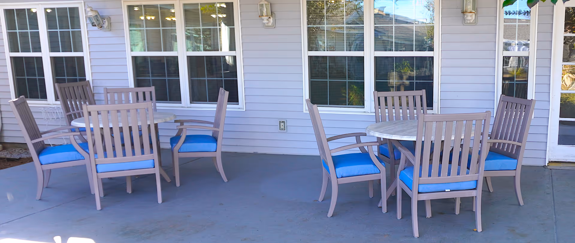 Outdoor patio area with two round tables surrounded by wooden chairs with blue cushions, situated in front of a building with white siding and multiple windows.