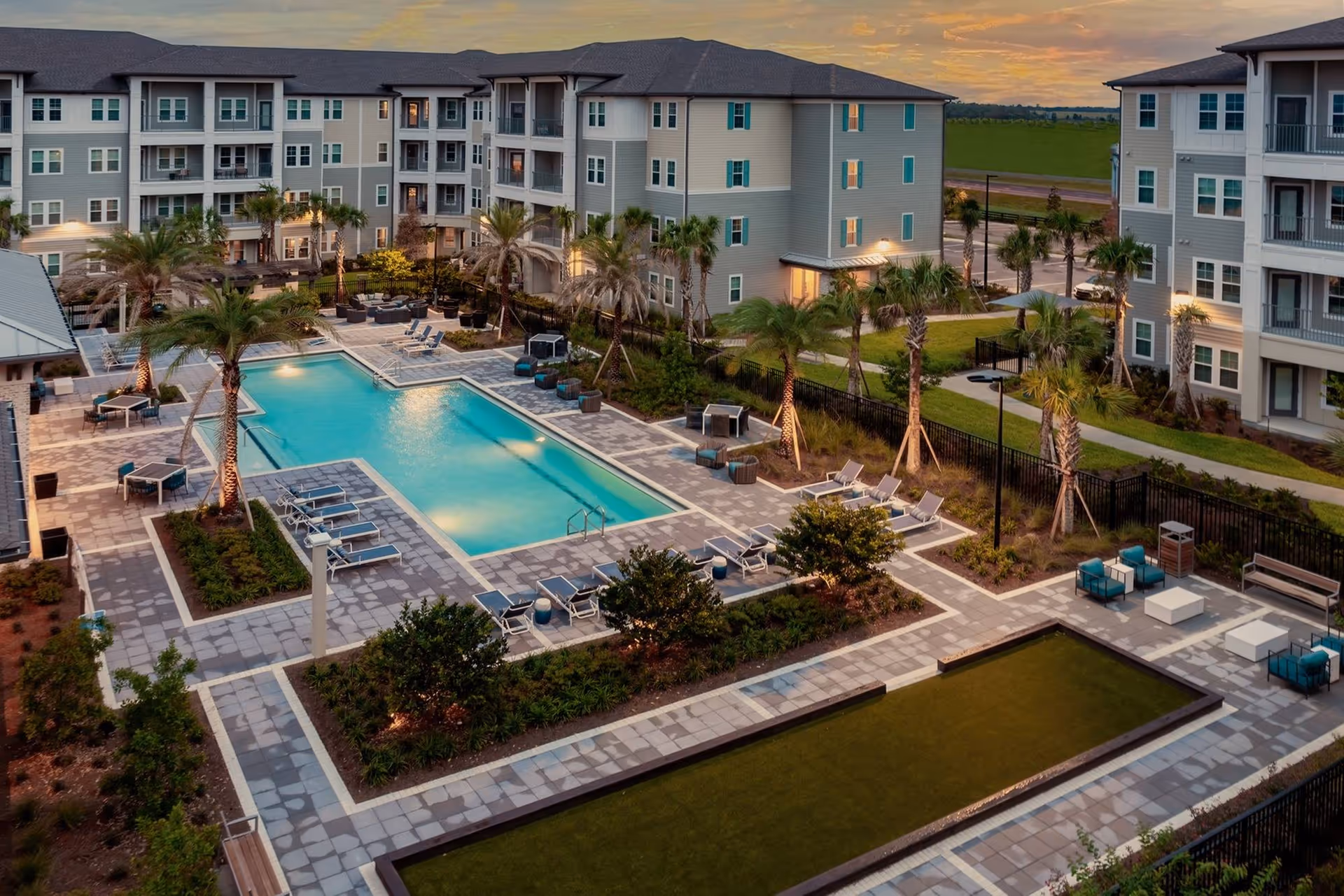 Aerial view of a landscaped courtyard with a swimming pool, lounge chairs, palm trees, and surrounding apartment buildings at sunset.