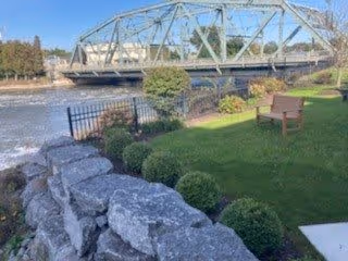 A peaceful outdoor scene featuring a metal truss bridge over a flowing river, with a stone wall and neatly trimmed bushes in the foreground. There is a grassy area with a wooden bench and a black metal fence along the riverbank.