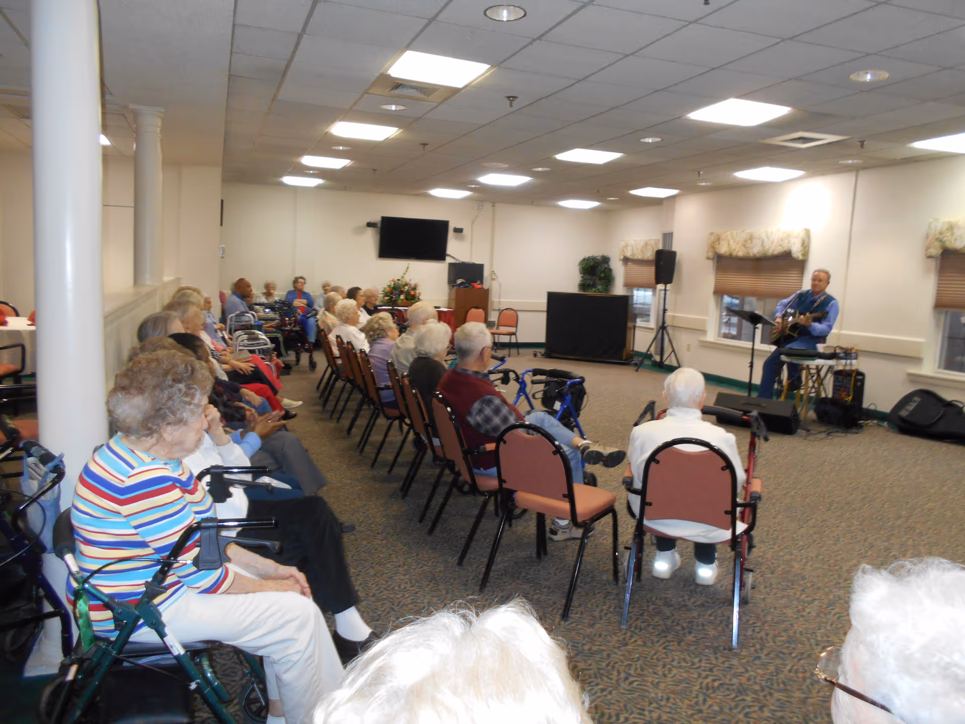 A group of elderly people seated in rows of chairs in a large room, attentively watching a man playing guitar and singing at the front. The room has carpeted floors, white walls, and ceiling lights. Some attendees have walkers and canes.