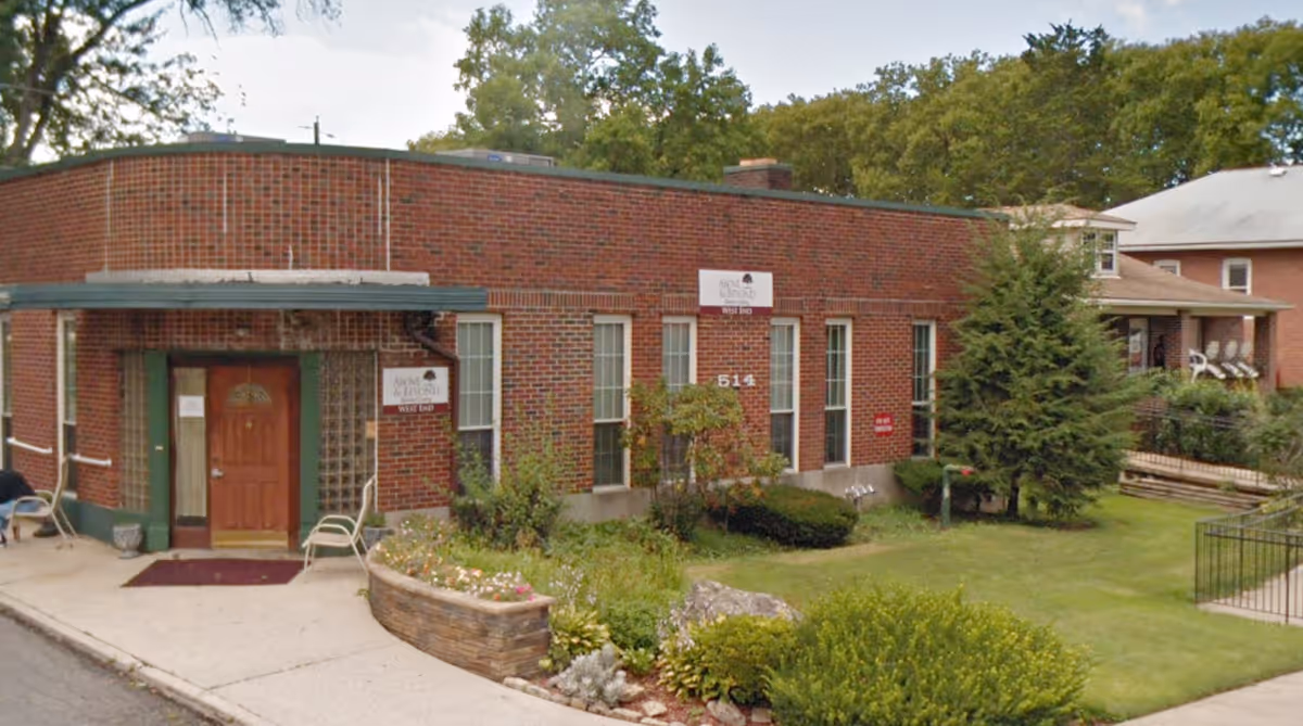 Exterior view of a single-story brick building with a green awning over the entrance door. There are several windows along the front wall, a small garden with various plants and shrubs, and a lawn area. A sign on the building reads 'West End'.