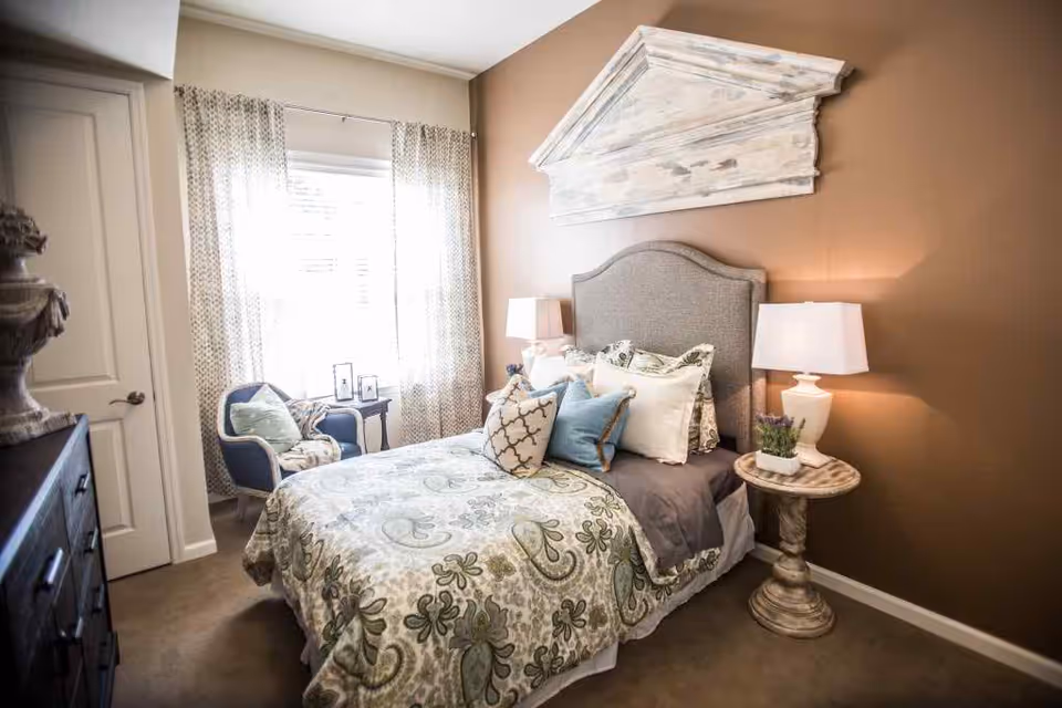 Cozy decorated bedroom with a bed dressed in patterned bedding, bedside tables with lamps, an armchair by a window, and wall decor above the headboard.