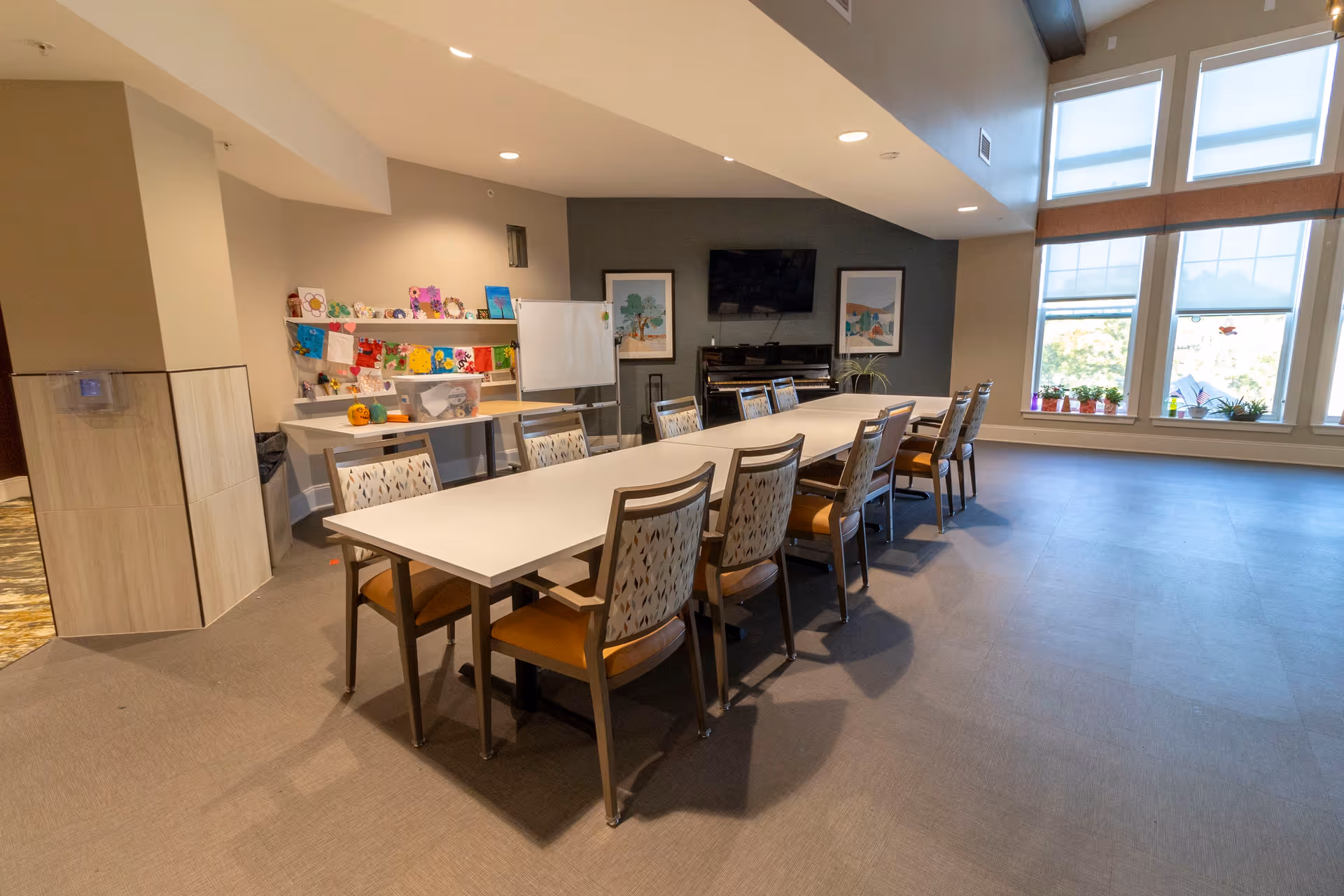 Long rectangular table surrounded by chairs in a bright communal activity/dining room with a TV, piano, craft supplies on a side table, and large windows.