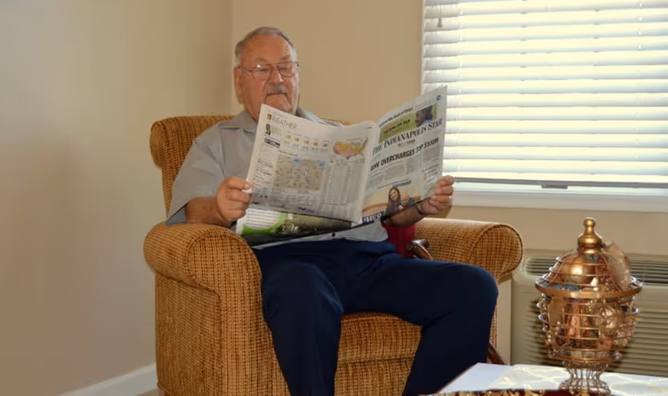 An elderly man sitting in a cushioned armchair reading a newspaper in a room with beige walls and a window with white blinds. There is a decorative copper item on a table nearby.