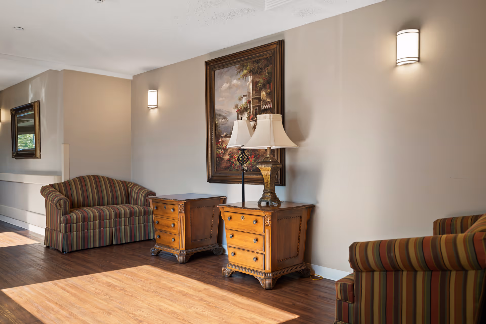 A cozy sitting area in a senior living facility hallway featuring two striped armchairs, two wooden side tables with drawers, a decorative table lamp, a framed painting on the wall, and wall-mounted light fixtures. The floor is wooden and sunlight streams in from the left side.