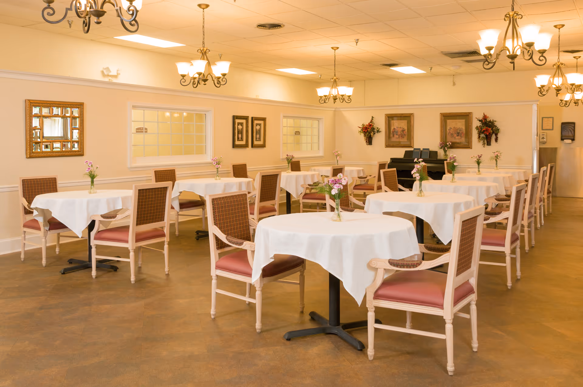 A dining room with multiple round tables covered with white tablecloths, each table having a small vase with flowers. The room has wooden chairs with cushioned seats, chandeliers hanging from the ceiling, framed artwork on the walls, and a piano in the background.