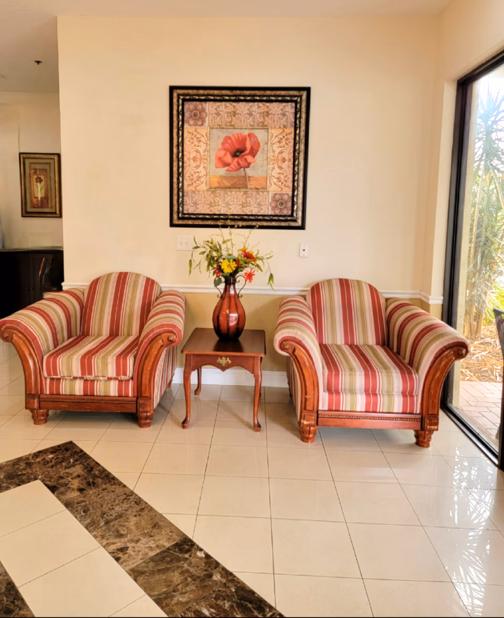 Two striped upholstered armchairs with wooden frames are placed on either side of a small wooden table holding a vase with flowers. Above the table hangs a framed painting of a red flower. The floor is tiled with a pattern of cream and brown tiles, and there is a large window on the right side showing outdoor plants.