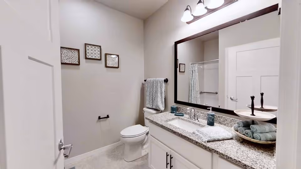 A clean and modern bathroom featuring a white toilet, a granite countertop with a sink, a large rectangular mirror with dark frame, and a three-light fixture above the mirror. The countertop holds neatly folded towels, soap dispensers, and a tiered tray with rolled towels. The walls are light-colored with three small framed artworks, and a towel hangs on a black towel bar next to the toilet. A shower with a glass door is visible in the mirror reflection.