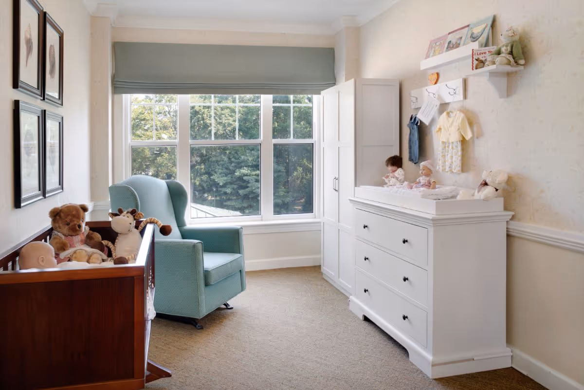 Sunlit nursery-style bedroom with a crib full of stuffed animals, a teal armchair, and a white dresser/changing table by a large window.