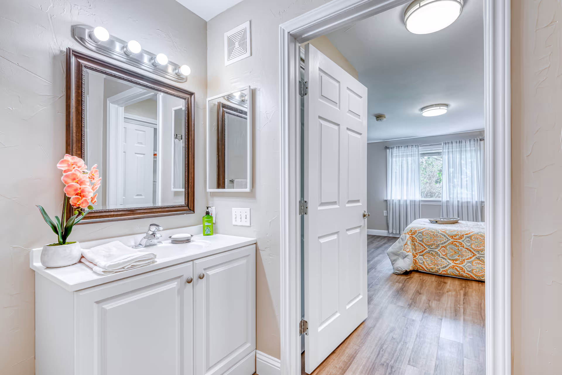 A bright bathroom vanity area with a white cabinet, a sink, a large framed mirror, and a smaller medicine cabinet. There is a pink orchid plant and folded white towels on the countertop. The bathroom opens into a bedroom with a bed covered in a patterned orange and gray comforter, wood flooring, and a window with sheer curtains.