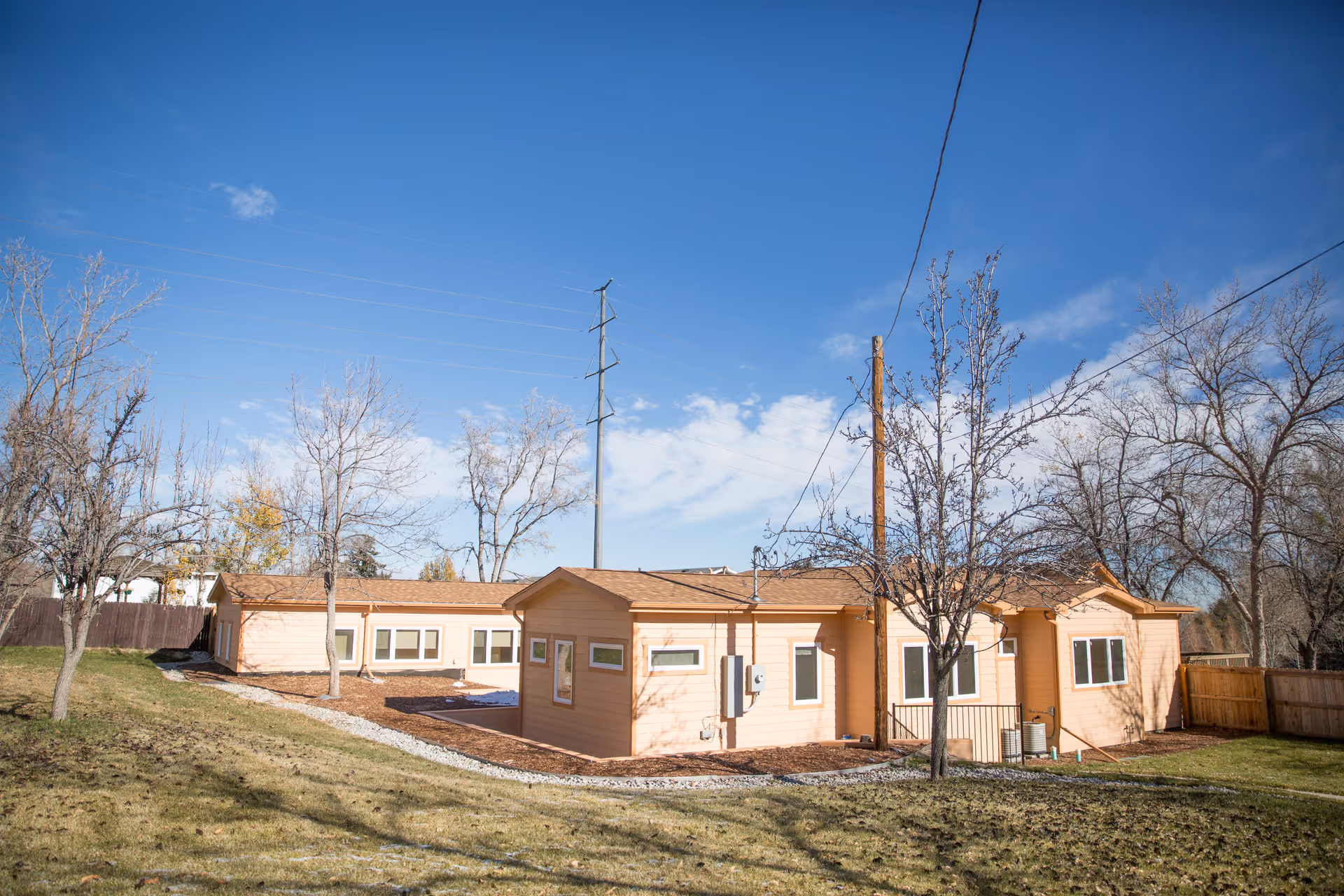 Exterior view of a single-story peach-colored building with multiple windows, surrounded by leafless trees and a grassy yard under a clear blue sky.
