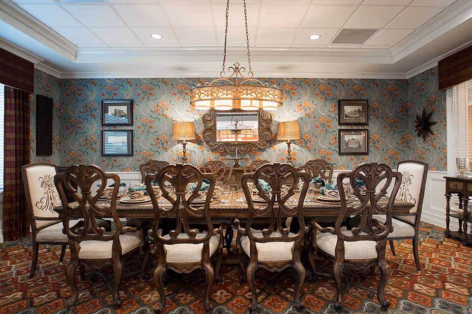 A formal dining room with a large wooden dining table surrounded by ten intricately carved wooden chairs with upholstered seats. The walls are decorated with floral patterned wallpaper and framed artwork. Two table lamps with beige shades flank a decorative mirror on the far wall. A large, ornate chandelier hangs above the table. The room has a patterned carpet and windows with plaid curtains.