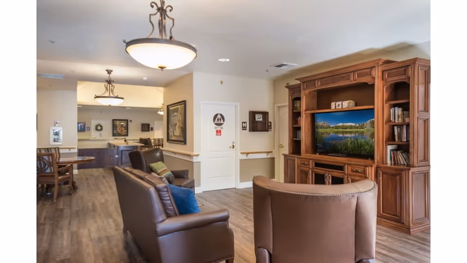 A cozy senior living common area with brown leather sofas and armchairs facing a large wooden entertainment center with a flat-screen TV displaying a nature scene. The room has wood flooring, soft lighting from ceiling fixtures, and a small dining area with tables and chairs in the background. The walls are light-colored with framed artwork and a door marked with restroom signs.