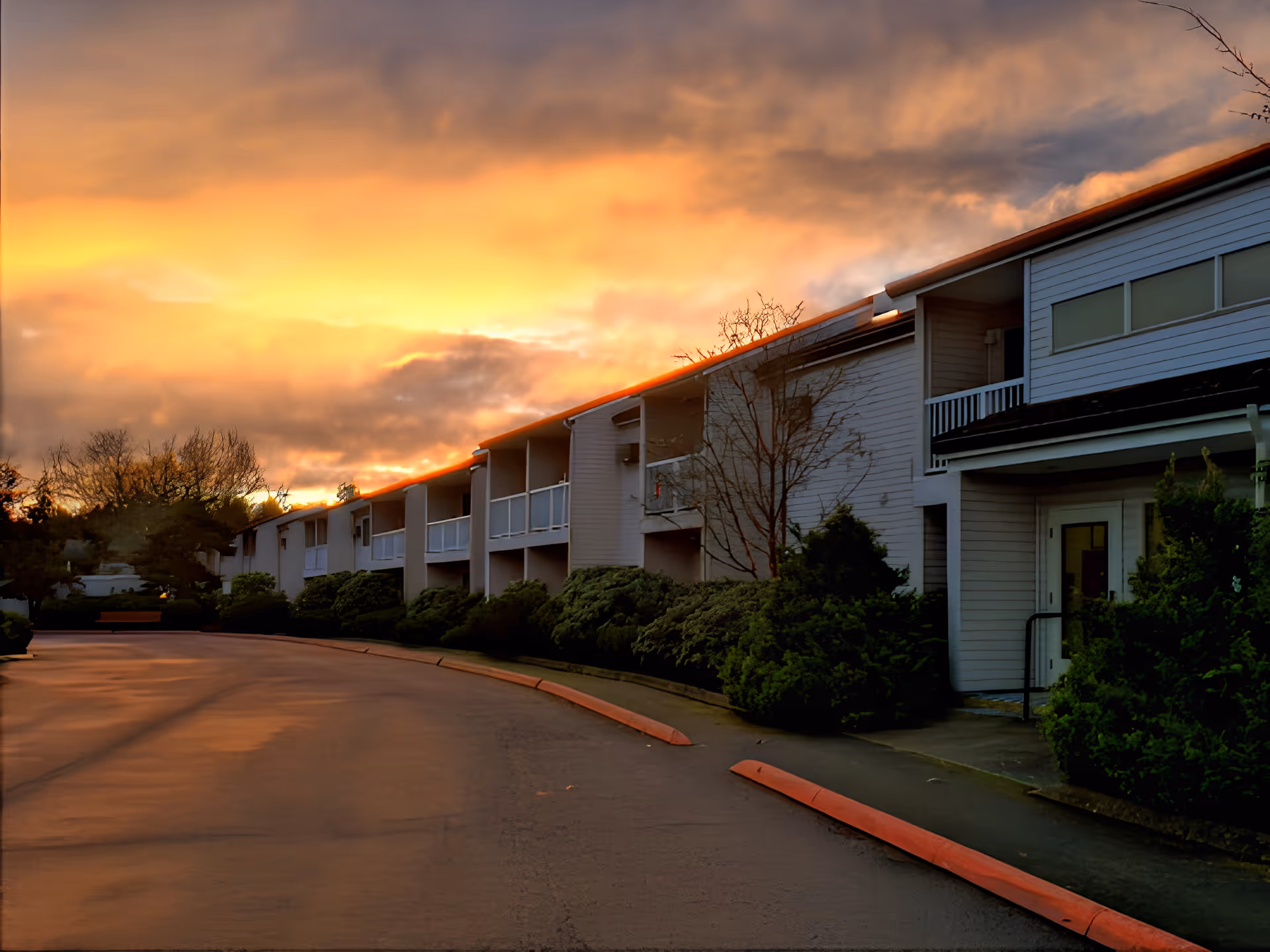 Exterior view of a retirement community building at sunset with a curved driveway, bushes lining the building, and a dramatic orange and yellow sky with clouds.