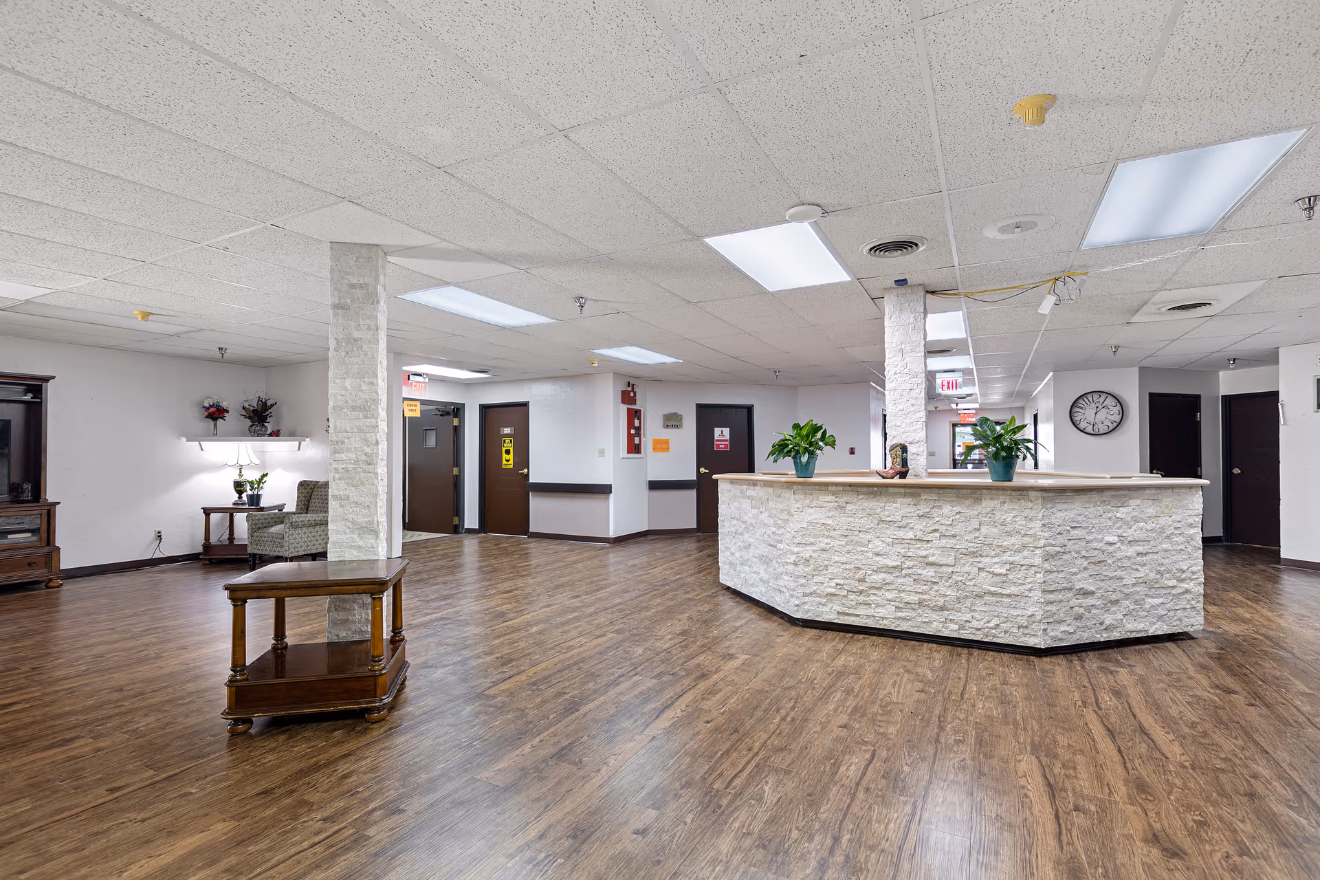 Interior view of a spacious reception area in a senior living facility with a large stone-faced reception desk, wooden flooring, white ceiling tiles with fluorescent lights, a small wooden table with plants, and several doors leading to other rooms.