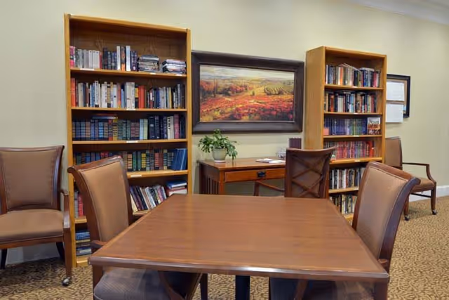 A cozy interior room with a wooden table surrounded by four cushioned chairs. Behind the table are two tall wooden bookshelves filled with books. A wooden desk with a potted plant and some papers sits between the bookshelves. Above the desk hangs a framed landscape painting with warm colors. The room has beige walls and carpeted flooring.