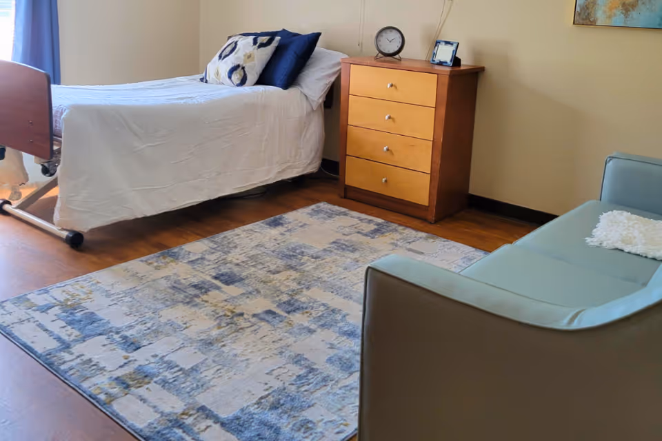 A senior living facility bedroom with a single bed covered in a white bedspread and two pillows, a wooden three-drawer dresser with a clock and photo frame on top, a patterned area rug on a wooden floor, and a light blue sofa with a white cushion.