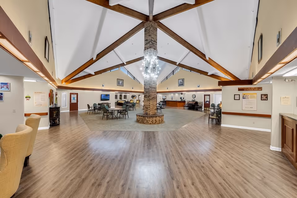 Spacious senior living facility common area with high vaulted ceilings featuring wooden beams and a central brick column with a modern chandelier. The room has wood flooring around a carpeted seating area with multiple tables and chairs. Walls are decorated with framed pictures and informational posters. There is a reception desk on the right side and a door on the left side.