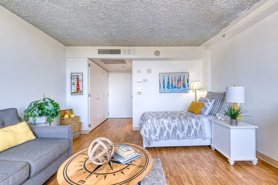 A bright and cozy senior living room featuring a gray sofa with a yellow pillow, a round wooden coffee table with decorative items and books, a single bed with patterned bedding and yellow accent pillows, a white nightstand with a lamp and plant, wooden flooring, and colorful wall art depicting sailboats and a lighthouse.