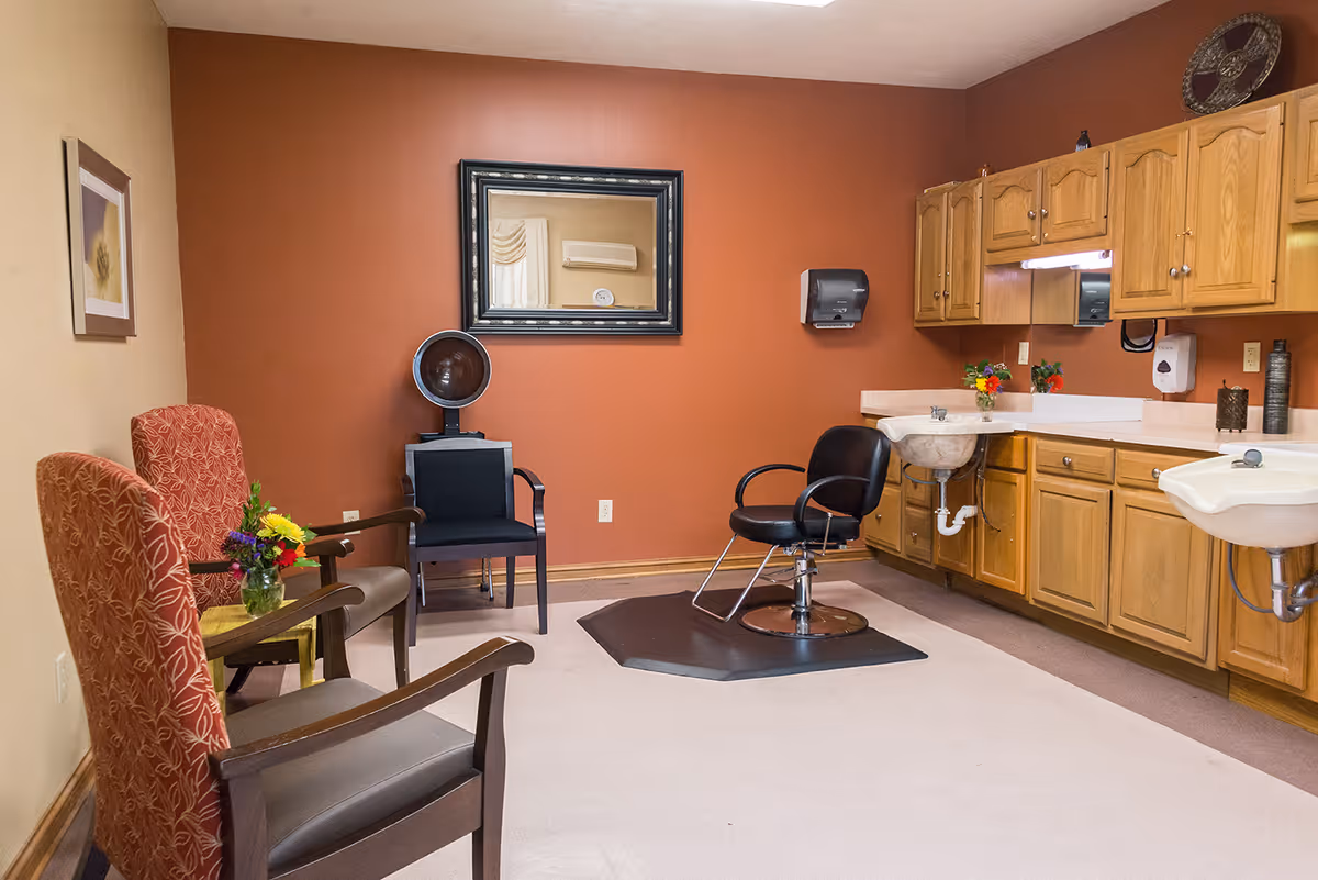 Interior salon-style room with a black stylist chair on a mat, hooded hair dryer, two sinks and wooden cabinets against a red accent wall, and guest chairs with a small table and flowers.