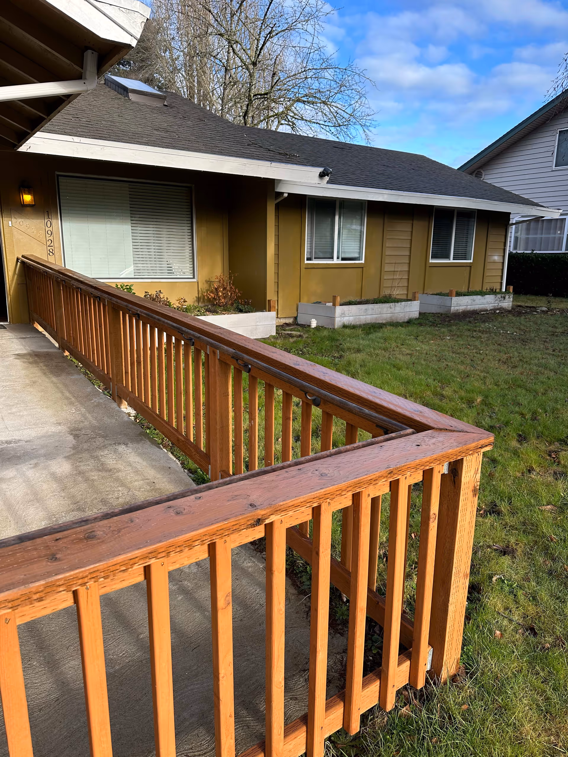 Exterior view of a single-story building with a wooden ramp leading to the entrance. The building has mustard yellow siding, three windows with white blinds, and a small garden area with raised planter boxes. The sky is partly cloudy and there is green grass surrounding the ramp.