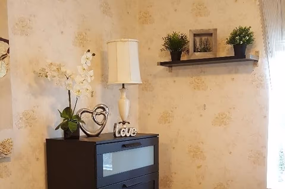 A decorative room corner with a lamp, potted plants, a heart sculpture and a small black dresser beneath a wall shelf.