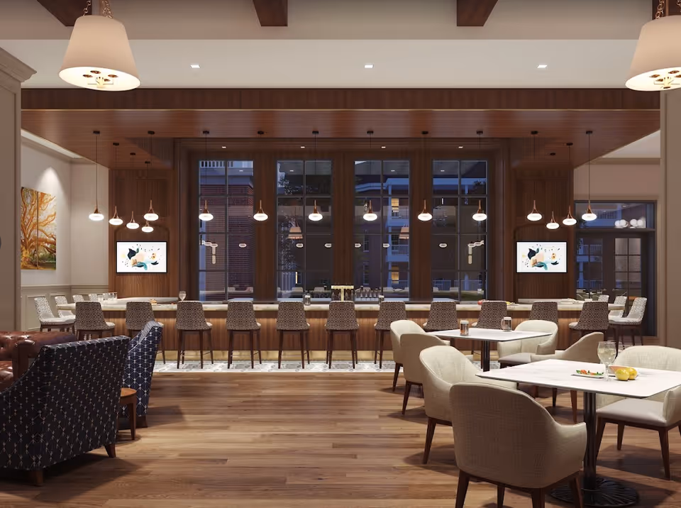 A spacious and modern dining area with a long bar counter lined with high chairs, pendant lights hanging above, and large windows in the background. The room features wooden flooring, comfortable upholstered chairs around tables, and decorative paintings on the walls.