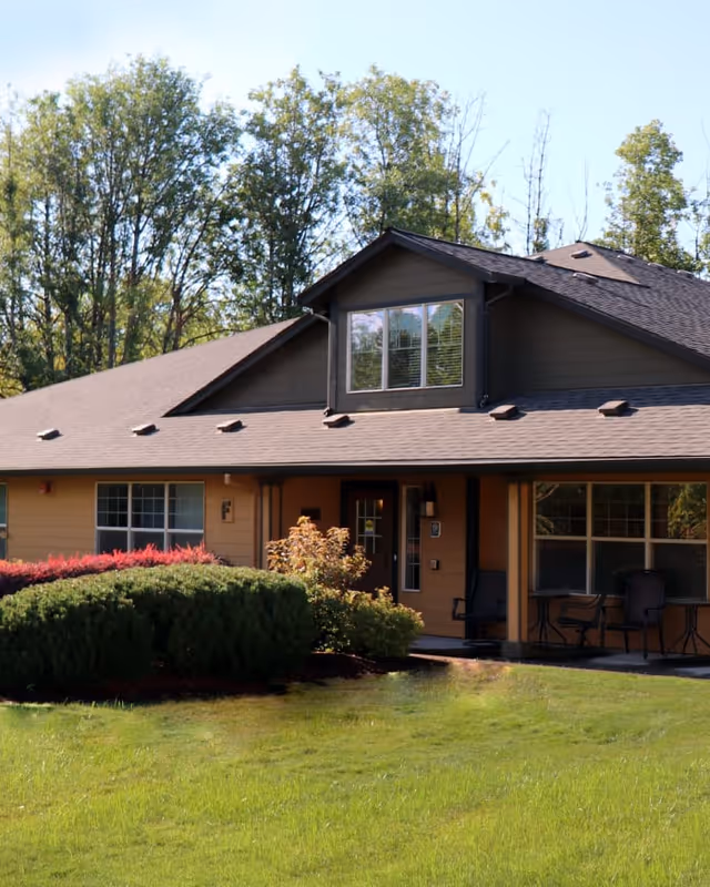 Front of a single-story residential-style building with a porch, lawn, shrubs, and trees in the background.