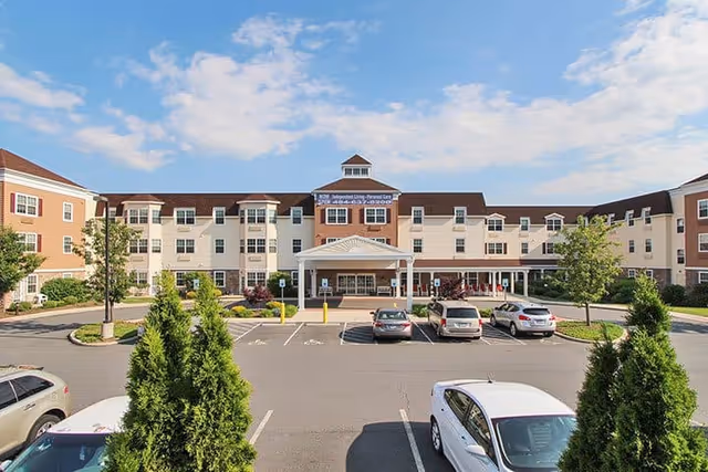 Front exterior view of Keystone Villa at Fleetwood, a multi-story senior living facility with a covered entrance, several parked cars in the parking lot, landscaped greenery, and a partly cloudy blue sky.