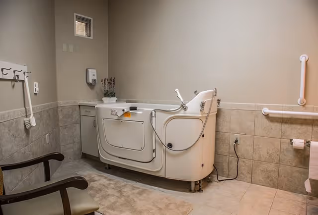 A bathroom designed for assisted living with a walk-in bathtub, beige tiled walls and floor, grab bars on the walls, a chair, a small cabinet with a plant, and a soap dispenser mounted on the wall.