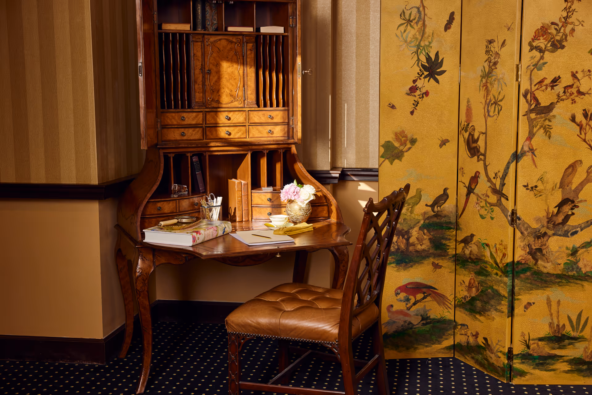 A vintage wooden writing desk with multiple small drawers and compartments, holding books, a cup with pens, a notebook, and a small vase with flowers. A brown leather cushioned chair is placed in front of the desk. To the right, there is a decorative folding screen with a painted design of birds and flowers. The room has beige walls with a striped pattern and a dark carpet with a dotted pattern.