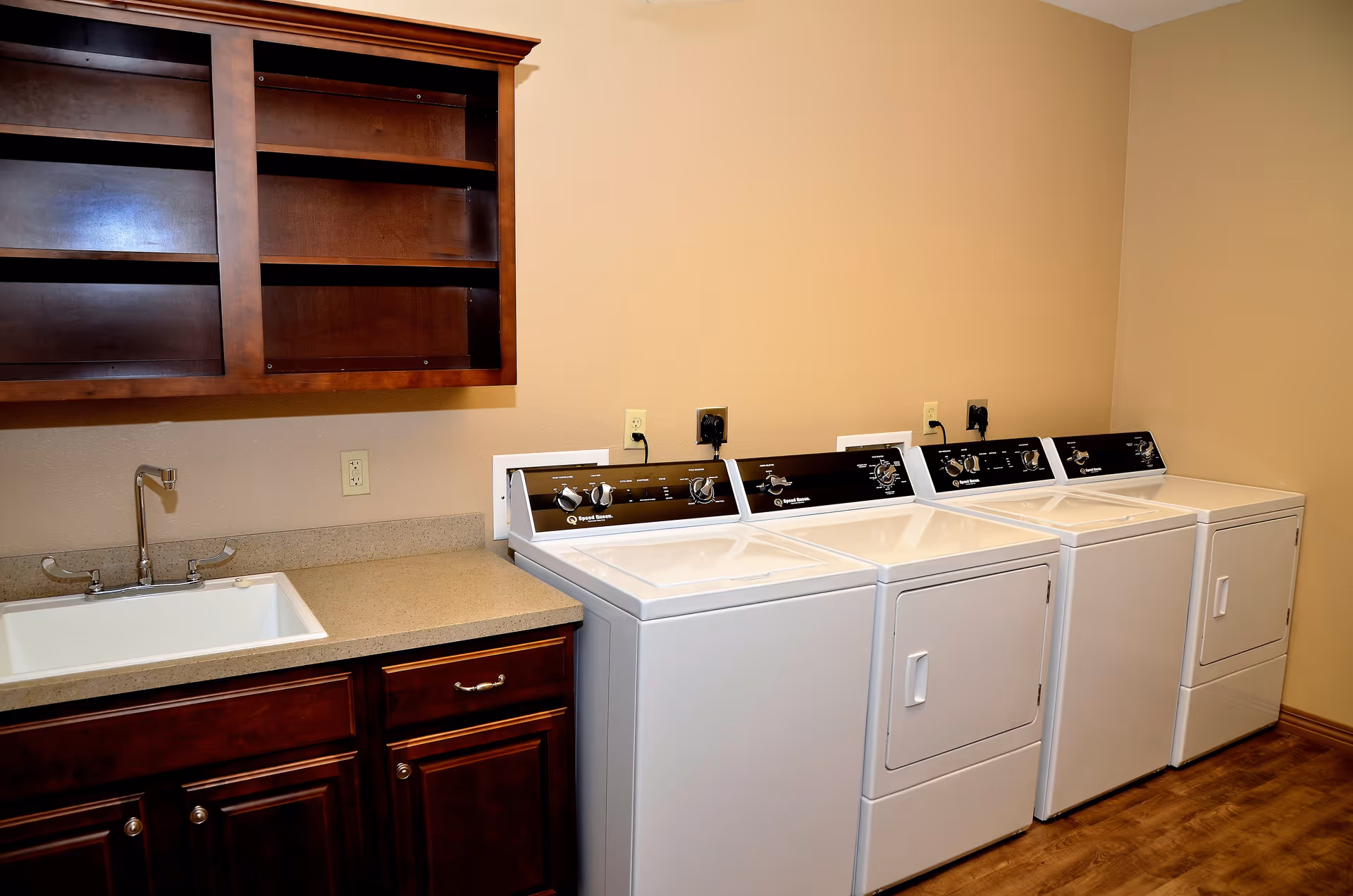 Laundry room with three white washing machines and dryers lined up against a beige wall, next to a countertop with a sink and dark wooden cabinets above and below.