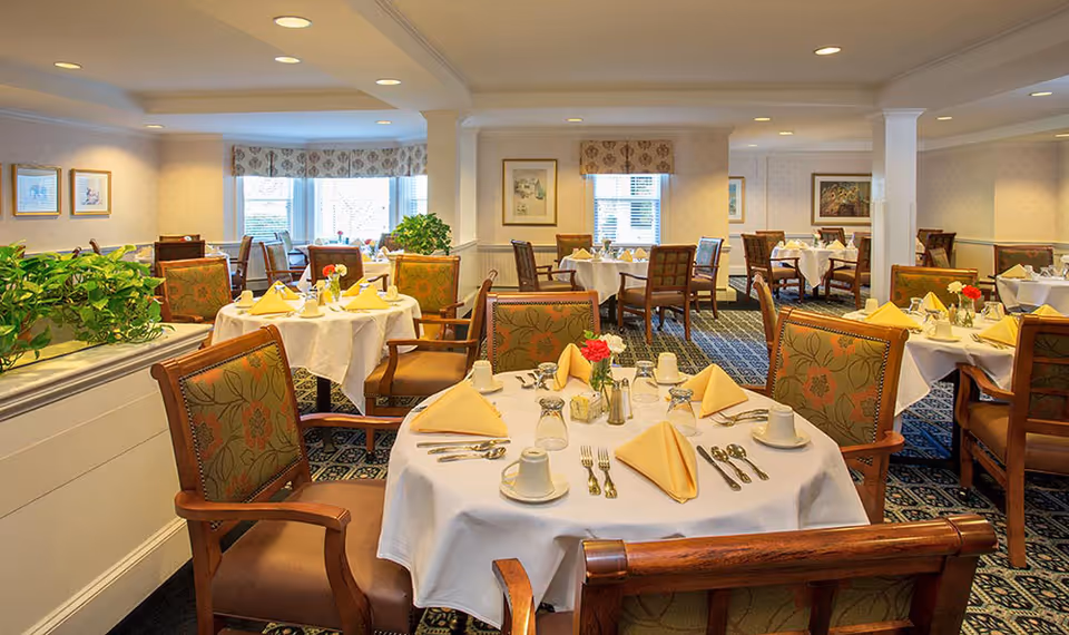 A well-lit dining room with multiple round tables covered with white tablecloths, each set with yellow folded napkins, cups, glasses, and silverware. The chairs have wooden frames with floral patterned upholstery. There are plants on a divider and framed artwork on the walls, with windows letting in natural light.